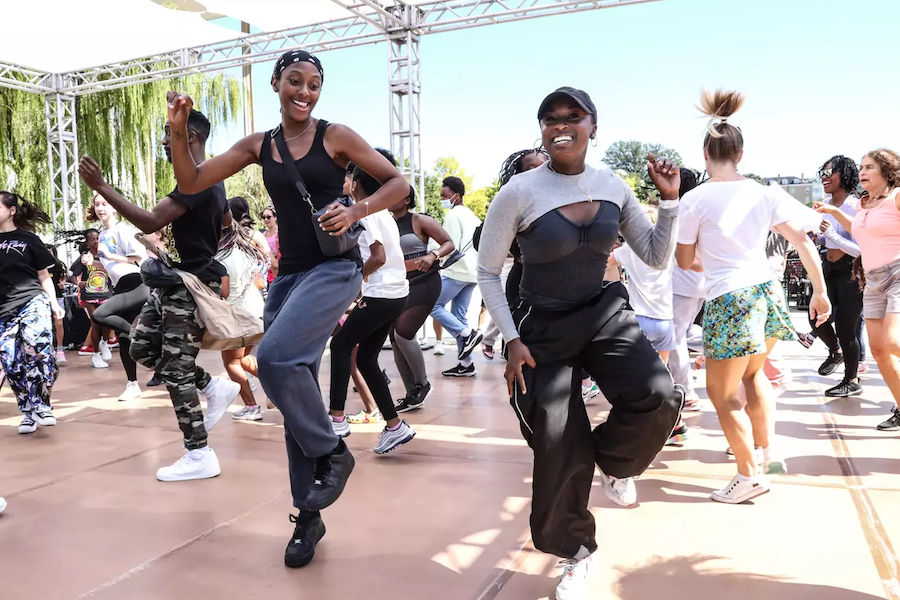 A group of people energetically dancing together outdoors under a canopy, led by two women in the foreground who are smiling and having fun.