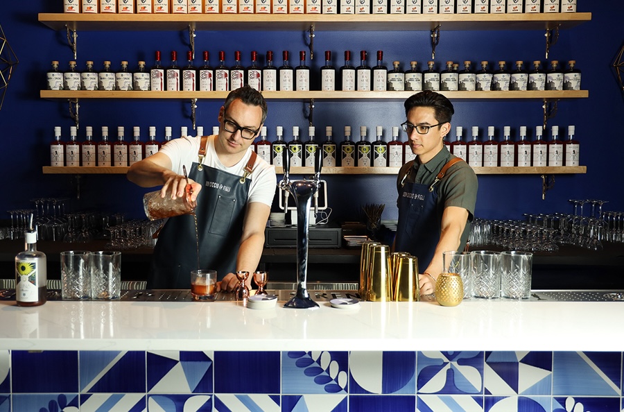 a male bartender with glasses and an apron pours a cocktail while another looks on, with shelves of liqueurs in the back