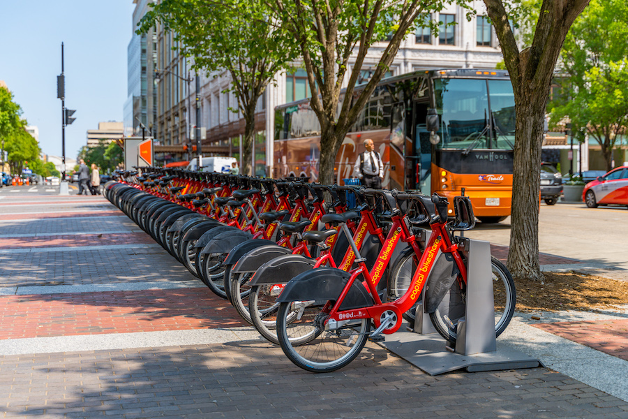 A row of red rental bicycles lined up at a docking station on a busy urban street with trees and a bus in the background.