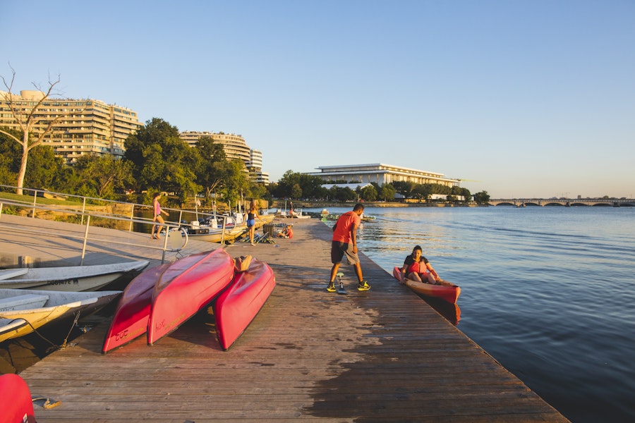People preparing to kayak on a waterfront dock with red kayaks, with buildings and trees in the background.