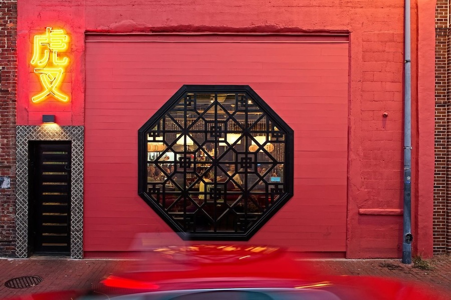 The exterior of Tiger Fork, a restaurant with a distinct red facade featuring an octagonal window and a bright yellow neon sign with Chinese characters. A blurred red car passes by in the foreground, adding a sense of movement to the scene.