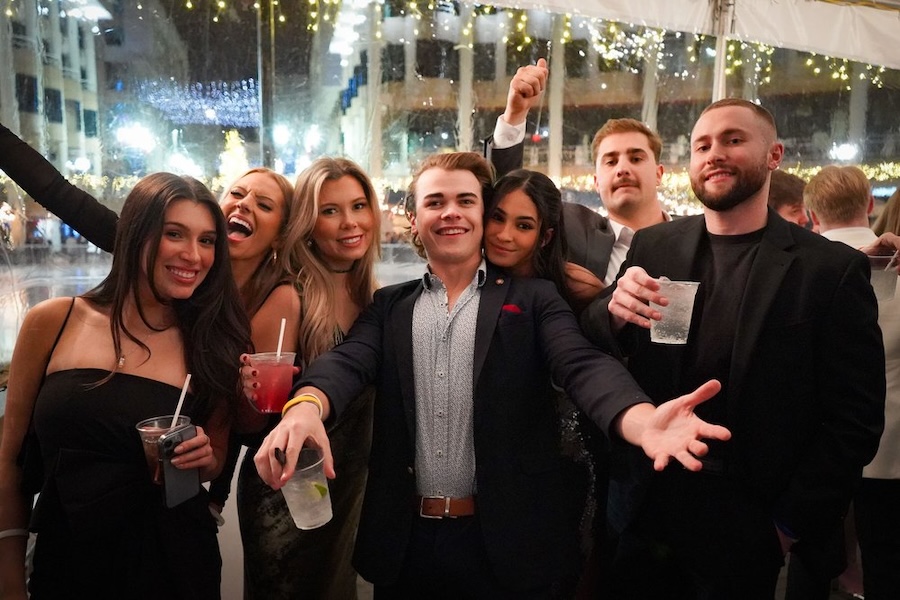 A group of smiling friends poses together at a festive event, enjoying drinks and celebrating in front of a backdrop of twinkling lights.