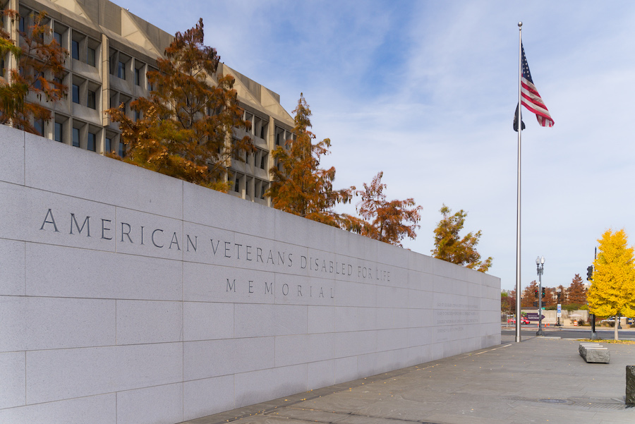 The American Veterans Disabled for Life Memorial features a stone wall engraved with its name, set against autumn trees and a flagpole flying the American flag.