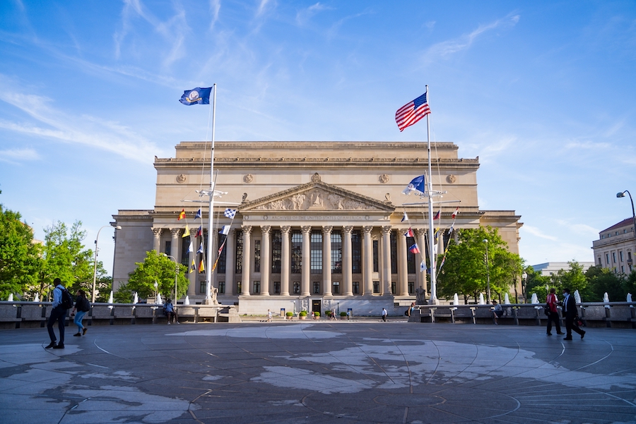 The National Archives building stands under a clear blue sky, framed by two flagpoles with American and military flags at the entrance, and a few people walking in the foreground.