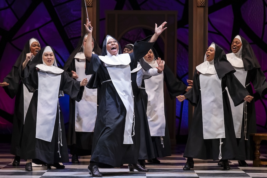 A nun holding a conductor ’ s wand dances animatedly with arms stretched up in the air as a large group of nuns dance and sing behind her.