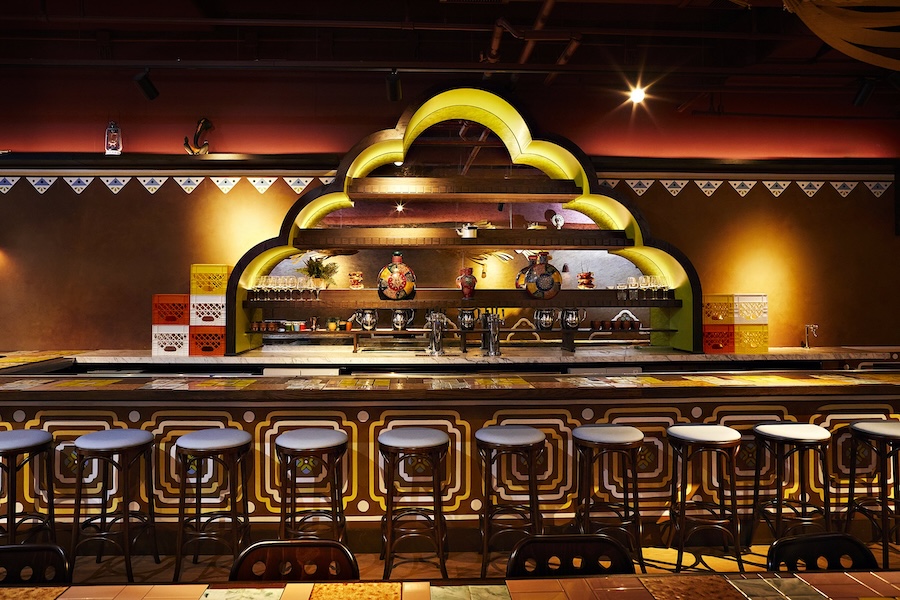 A vibrant, ornately decorated bar with colorful tiles, patterned stools and a backlit arch-shaped shelf displaying glassware and decorative pottery.